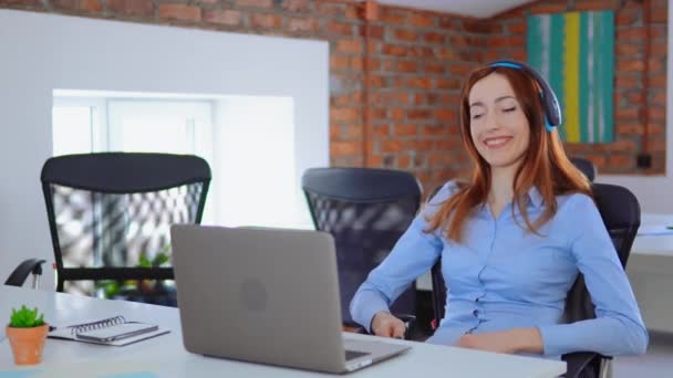 Lycklig ung kvinna sjunger medan du lyssnar på hög musik i hörlurar sitter på kontoret skrivbord med laptop — Stockvideo