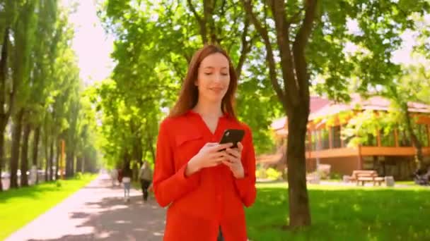 Pretty red-haired woman walking and texting messages in park — Stock Video