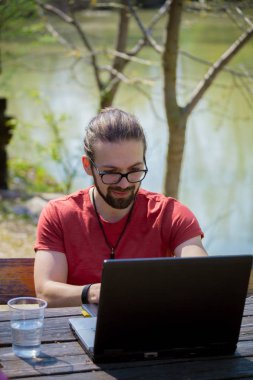 Genç, gülümseyen adam, serbest tasarımcı, göl kenarında çalışıyor.