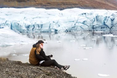 two tourists are sitting near the glacier iceberg in Iceland clipart