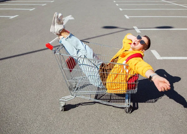 时尚的家伙在太阳镜和黄色夹克坐在购物车从食品在超市停车场 — 图库照片