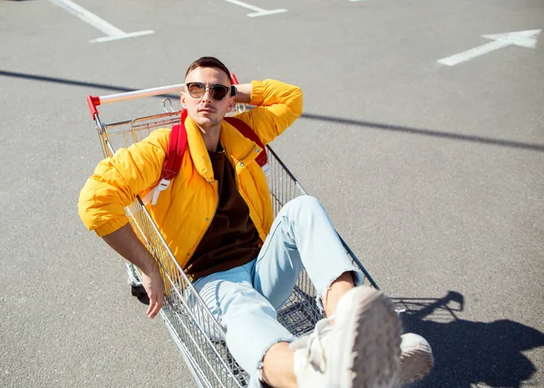 时尚的家伙在太阳镜和黄色夹克坐在购物车从食品在超市停车场 — 图库照片