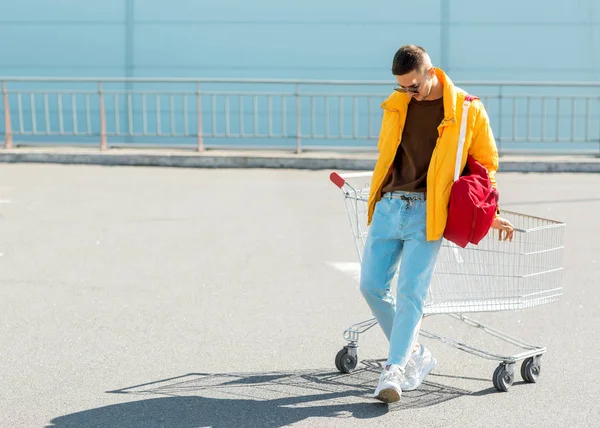时尚的家伙在太阳镜和黄色夹克跳在购物车从食品在超市停车场 — 图库照片