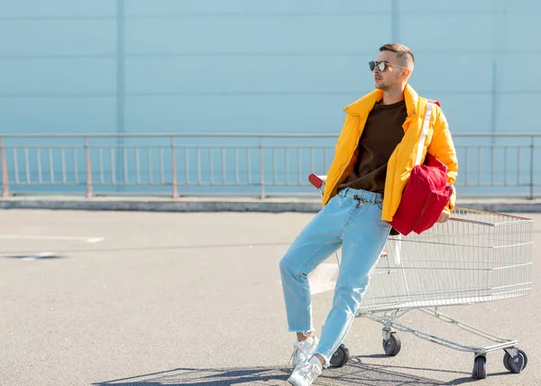 时尚的家伙在太阳镜和黄色夹克跳在购物车从食品在超市停车场 — 图库照片