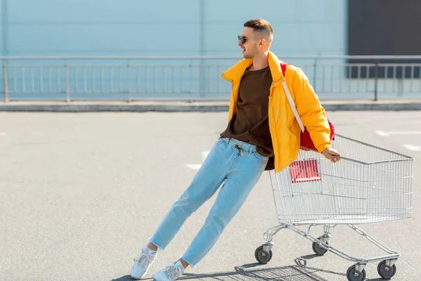 ファッションの男のサングラスと黄色のジャケットのスーパー マーケット駐車場で食物からカートにジャンプします — ストック写真