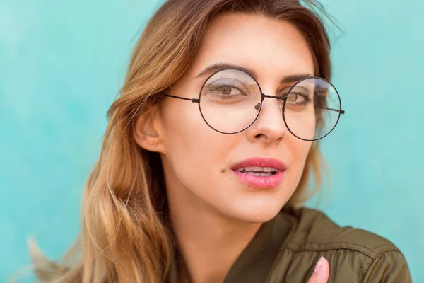 fashion girl in round glasses stands posing near a turquoise wall