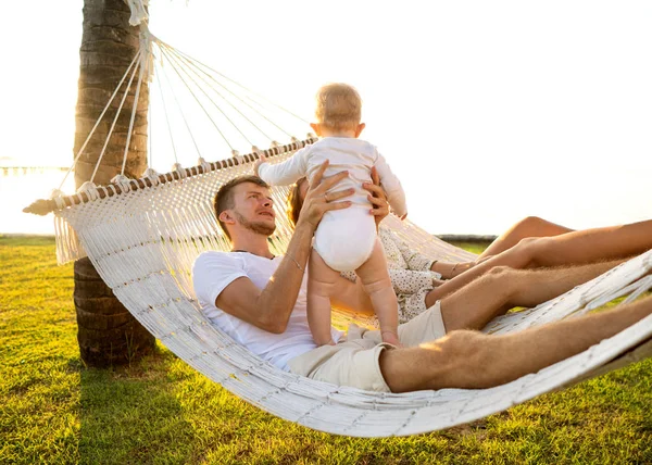 Família feliz em uma ilha tropical ao pôr do sol mentira em uma rede e brincar com seu filho — Fotografia de Stock