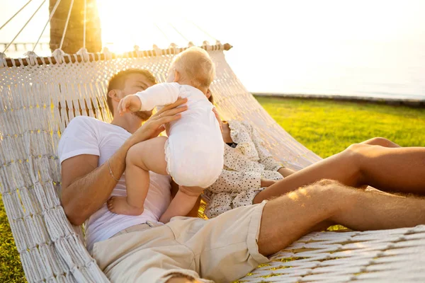 Família feliz em uma ilha tropical ao pôr do sol mentira em uma rede e brincar com seu filho — Fotografia de Stock