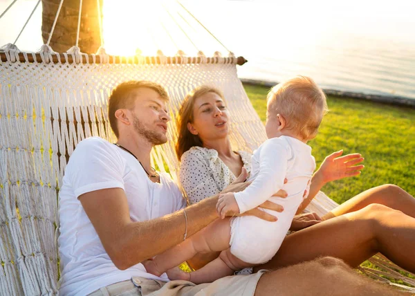 Família feliz em uma ilha tropical ao pôr do sol mentira em uma rede e brincar com seu filho — Fotografia de Stock