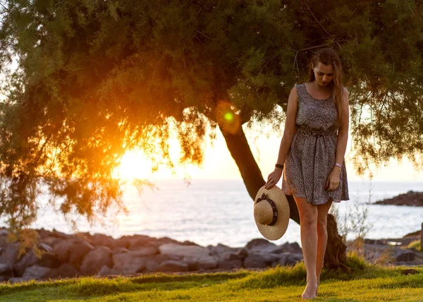 Schöne Mädchen mit Hut spaziert bei Sonnenuntergang in einem Kleid an der Küste entlang — Stockfoto