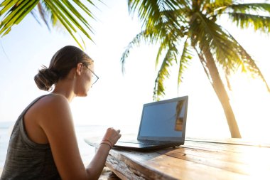 Tropikal palmiye ağaçları arasında bilgisayarı olan serbest çalışan kız gün batımında adada çalışıyor.