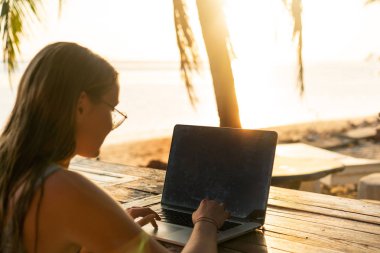 Tropikal palmiye ağaçları arasında bilgisayarı olan serbest çalışan kız gün batımında adada çalışıyor.