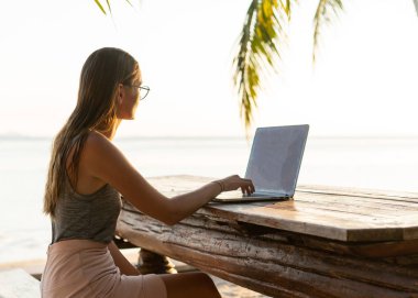 Tropikal palmiye ağaçları arasında bilgisayarı olan serbest çalışan kız gün batımında adada çalışıyor.