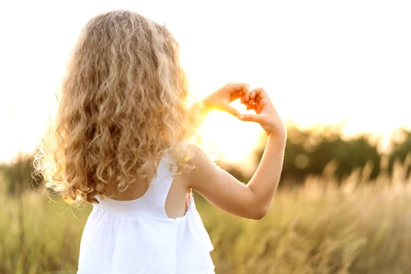 Klein meisje bij zonsondergang toont een hart met handen speelt in het veld — Stockfoto