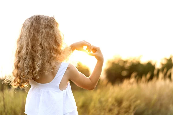 Menina ao pôr do sol mostra um coração com as mãos joga no campo — Fotografia de Stock