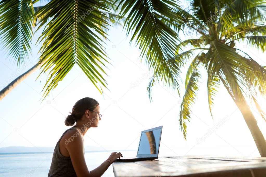 freelancer girl with a computer among tropical palm trees work on the island in sunset