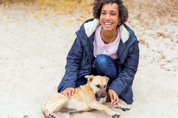 Sonbahar parkında güzel Afrikalı bir kız bir köpekle oynuyor. — Stok fotoğraf