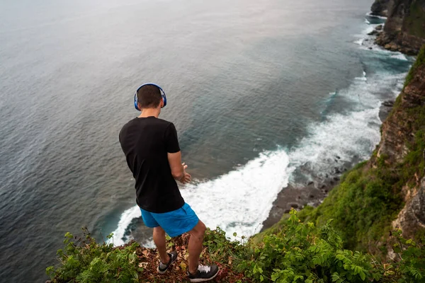 guy in headphones stands on a cliff by the ocean at sunset in bali