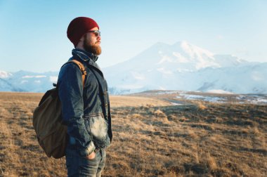 Düşünceli bir şık hipster güneş gözlüğü sakallı ve gün batımında Kafkasya'da karla kaplı Elbruz Dağı arka plan üzerinde bir sırt çantası ile bir şapka portre