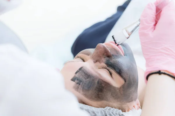 Kosmetyczki w różowe rękawiczki z pędzlem stosuje maski węgla dla peeling na twarzy młodej dziewczyny w Sala kosmetologii. Pojęcie usług kosmetologii i samoopieki. Pojęcie — Zdjęcie stockowe