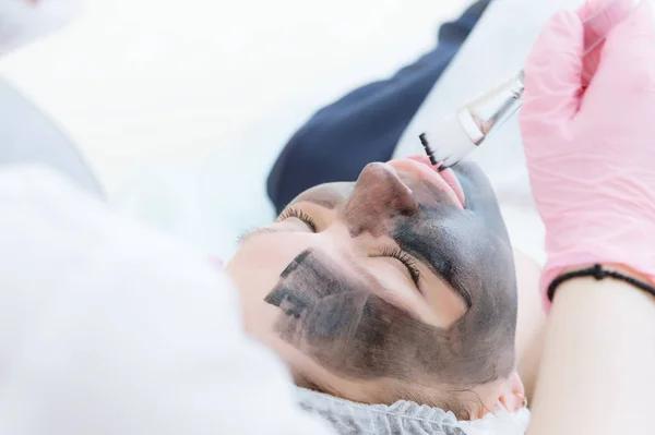 The cosmetologist in pink gloves with a brush applies a carbon mask for peeling on the face of a young girl in a cosmetology room. The concept of cosmetology services and self-care. The concept of