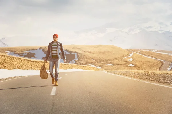 Un elegante hipster barbuto in occhiali da sole con uno zaino vintage cammina lungo la strada asfaltata in una giornata di sole. Il concetto di autostop e trekking — Foto Stock