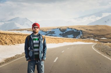 Güneş gözlüğü vintage bir sırt çantası ile şık bir sakallı hippi güneşli bir günde bir ülke yol asfalt üzerinde duruyor. Otostop ve hiking kavramı