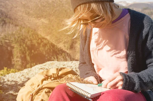 선글라스와 푸른 하늘에 대하여 산에 바위 야외에 앉아 모자를 입고 hipster 여자의 초상화. 여행에 프리랜서 디자이너 노트북에 크레용 파스텔을 그립니다. — 스톡 사진
