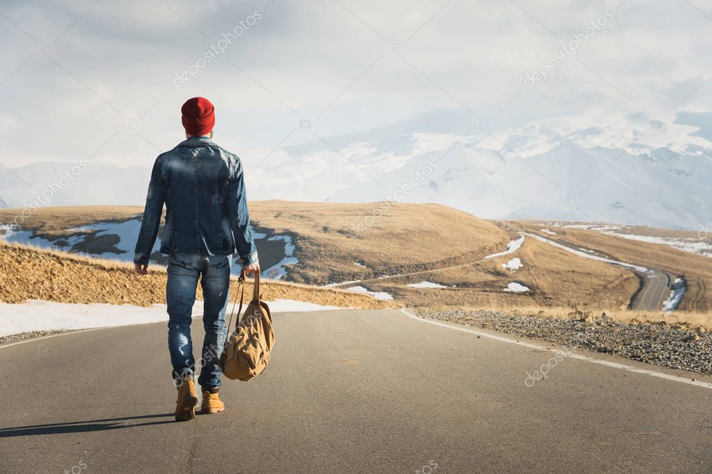 A stylish bearded hipster in sunglasses with a vintage backpack walks along the asphalt road on a sunny day. The concept of hitchhiking and hiking