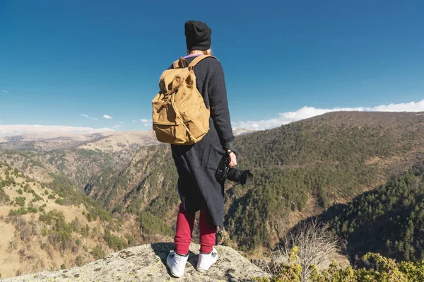 Een hipster reizen fotograaf meisje in een hoed en met een rugzak staande op een rots en kijken naar de heuvels. — Stockfoto