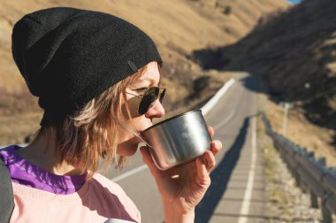 Güneş gözlüğü ve siyah bir şapka giyen bir hipster kız bir kupa bir termos içinde belgili tanımlık vahşi bir yolculuğa üzerinden gelen çay veya kahve içiyor