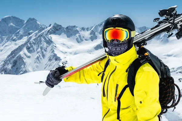 Un grande ritratto di uno sciatore con casco protettivo e occhiali è una maschera e una sciarpa con gli sci sulla spalla tra le montagne innevate del Caucaso. Sciare , — Foto Stock