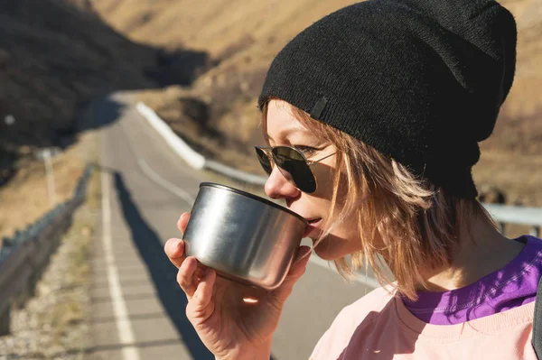 Uma garota hipster usando óculos de sol e um chapéu preto está bebendo chá ou café de uma caneca de uma garrafa térmica na natureza em uma viagem — Fotografia de Stock