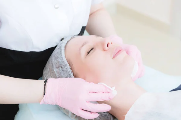 Um close-up do procedimento de limpeza no escritório de cosmetologia. As mãos do cosmetologista em luvas rosa são removidas do rosto de uma jovem com uma esponja limpando a máscara. — Fotografia de Stock