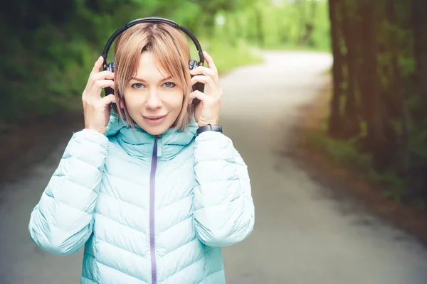 Portret Sporty atrakcyjny dziewczynka Blondynka w świetle działa w dół kurtka opatrunku bluetooth słuchawki z muzyką lub dźwięki natury na leśnej drodze — Zdjęcie stockowe