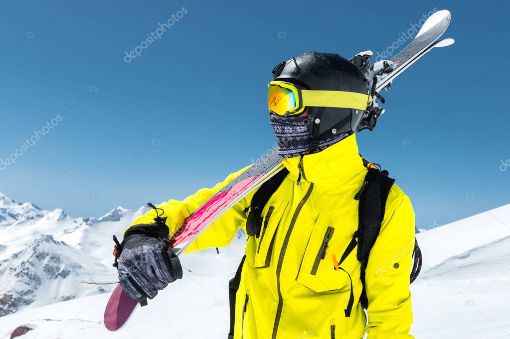 A large portrait of a skier in a protective helmet and glasses is a mask and scarf with skis on his shoulder in the snow-capped mountains of the Caucasus. Skiing,