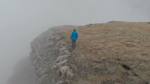 Hiker man in a blue jacket with backpack is walking and standing on the steep edge of mountain plateau above the clouds. Aerial view. Drone is orbiting. — Stock Video