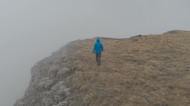 Man van de wandelaar in een blauwe jas met rugzak loopt en staande op de steile rand van het hoogplateau boven de wolken. Luchtfoto. Drone is een baan. — Stockvideo