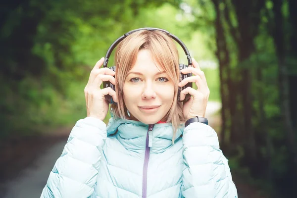 Portret Sporty atrakcyjny dziewczynka Blondynka w świetle działa w dół kurtka opatrunku bluetooth słuchawki z muzyką lub dźwięki natury na leśnej drodze — Zdjęcie stockowe