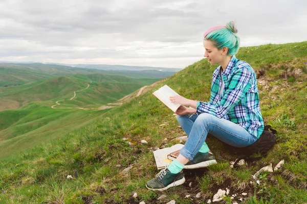 Retrato de uma jovem hipster com cabelos multicoloridos sentados na natureza nas montanhas lendo um livro. O conceito de leitura na natureza. Educação — Fotografia de Stock