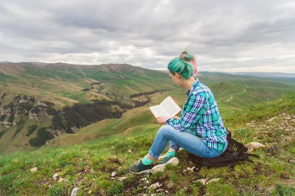 Retrato de uma jovem hipster com cabelos multicoloridos sentados na natureza nas montanhas lendo um livro. O conceito de leitura na natureza. Educação — Fotografia de Stock