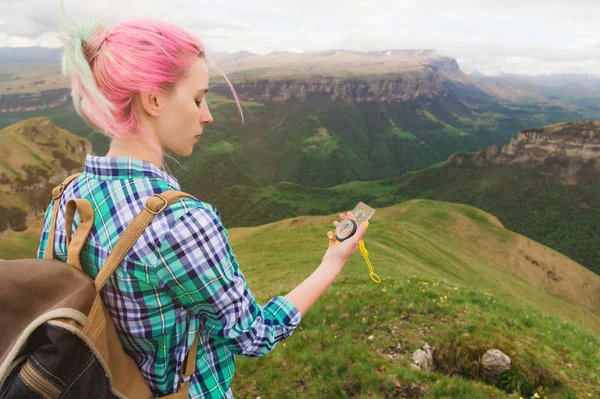 Uma garota hipster viajou com um blogueiro em uma camisa xadrez e com cabelo multicolorido usando uma bússola no fundo no fundo da paisagem caucasiana com um planalto — Fotografia de Stock