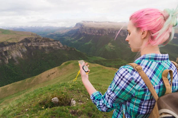 Uma garota hipster viajou com um blogueiro em uma camisa xadrez e com cabelo multicolorido usando uma bússola no fundo no fundo da paisagem caucasiana com um planalto — Fotografia de Stock