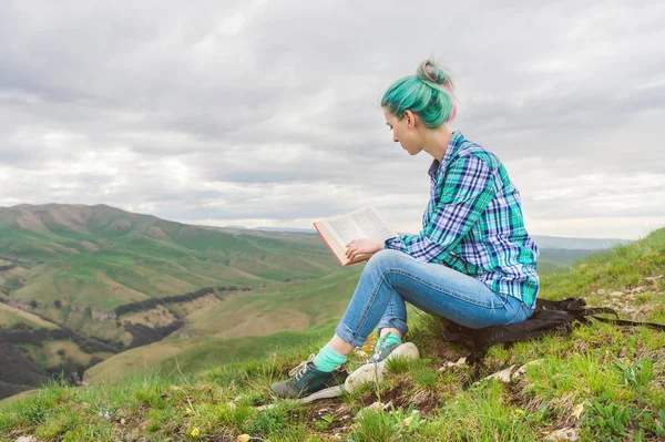 Retrato de uma jovem hipster com cabelos multicoloridos sentados na natureza nas montanhas lendo um livro. O conceito de leitura na natureza. Educação — Fotografia de Stock