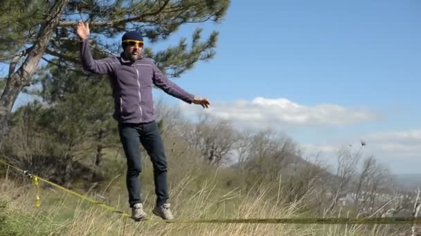 Een man die het dragen van een hoed en een baard loopt de slackline tijdens de wind tegen de achtergrond van de bergen van de Kaukasus van de blauwe lucht en de wolken — Stockvideo