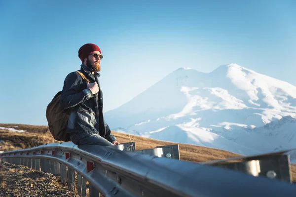 Barbuto turista hipster uomo in occhiali da sole con uno zaino seduto su un dosso stradale e guardando il tramonto sullo sfondo di una montagna innevata — Foto Stock