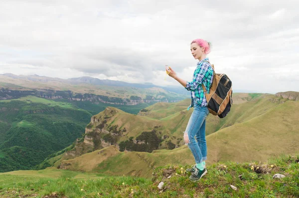 Uma garota hipster viajou por um blogueiro em uma camisa xadrez e com cabelo multicolorido usando uma bússola em pé em um fundo de platô nas montanhas — Fotografia de Stock