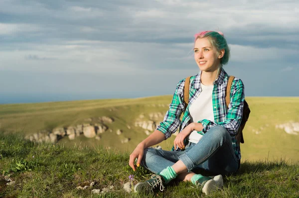 Retrato de jovem mulher sorridente viajante com cabelo multi-colorido. Sentado no alto das montanhas à noite com um declínio no fundo de um planalto — Fotografia de Stock