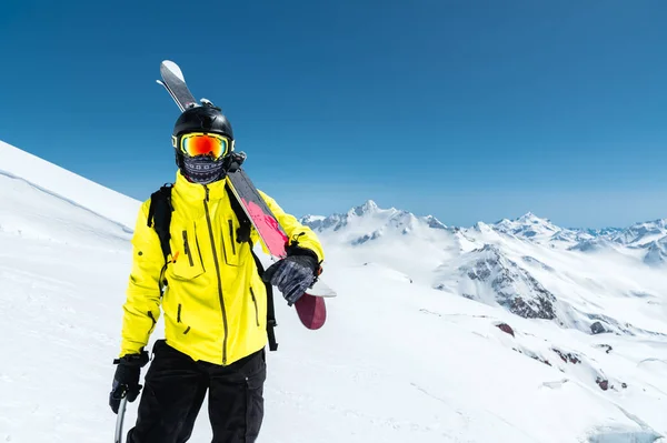 Ett porträtt av en skidåkare i en skyddande hjälm och glasögon är en mask och halsduk med skidorna på axeln i de snöklädda bergen i Kaukasus. Skidåkning, — Stockfoto