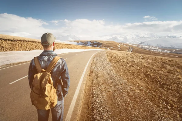 Un hombre barbudo con una gorra y una mochila lista para recorrer un largo camino. Un hombre en un camino rural contra el telón de fondo de montañas y nubes — Foto de Stock